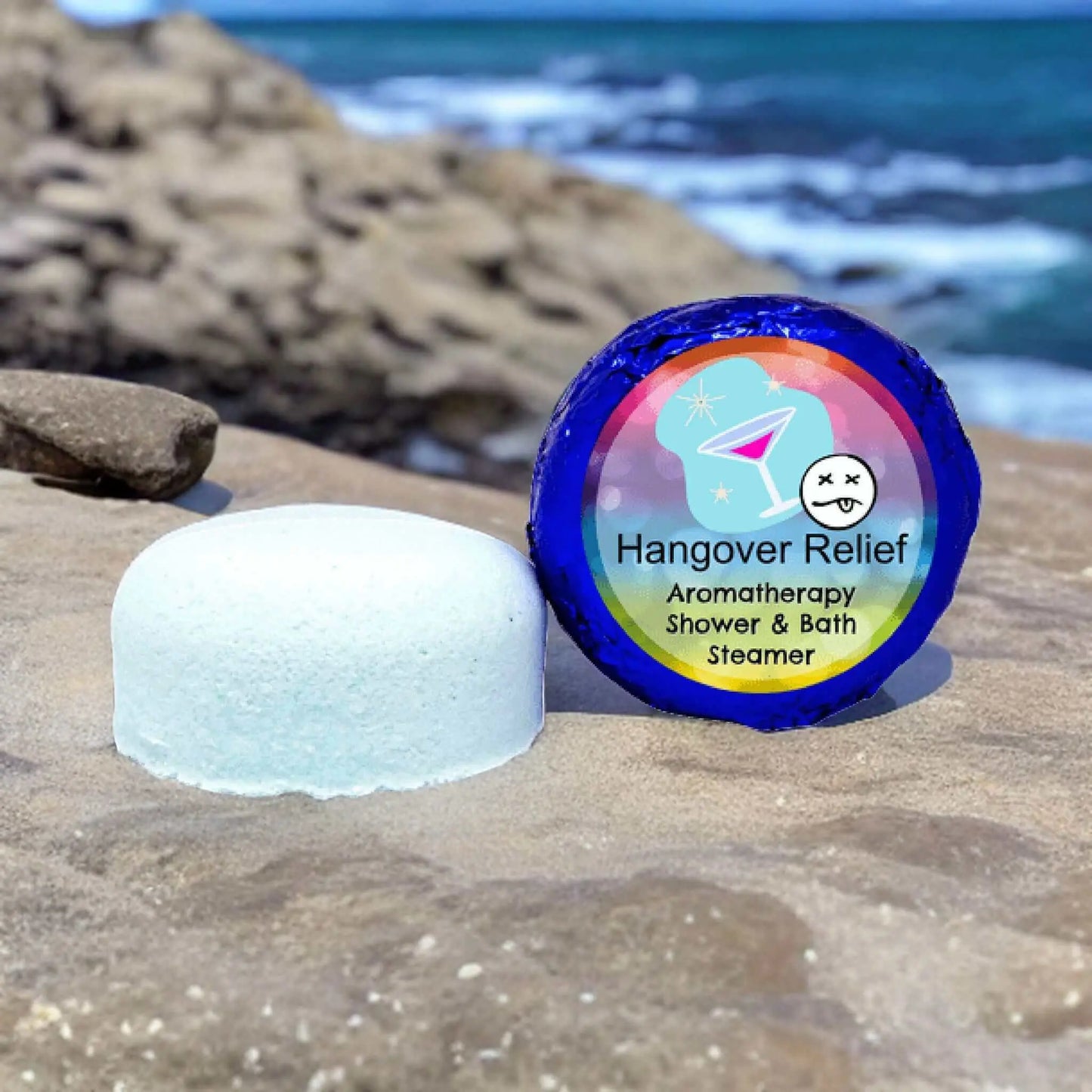 A round, white Posh Brats Hangover Relief Aromatherapy Shower Steamer sits on a rocky shore. Its colorful packaging, adorned with a cartoon cocktail, hints at its purpose. Infused with essential oils, it provides solace as ocean waves crash nearby.