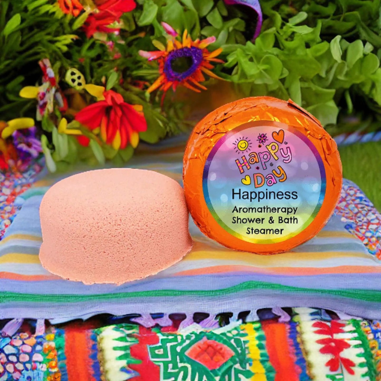 A round pink shower steamer labeled Happiness Aromatherapy Shower Steamer by Posh Brats sits on a striped cloth, with citrus aroma emanating from its colorful packaging. Vibrant flowers in the background enhance the cheerful setting with their essential oils allure.