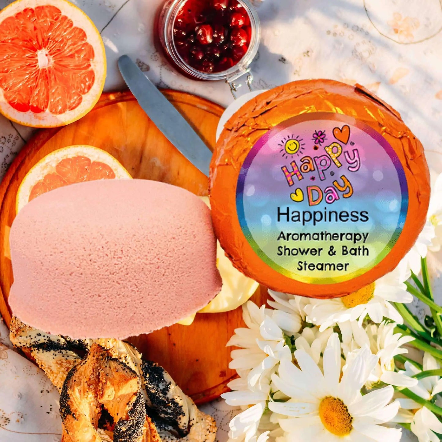 A vibrant flat lay features Posh Brats Happiness Aromatherapy Shower Steamer with citrusy essential oils, next to a pink soap. The scene includes sliced grapefruit, a jar of pomegranate seeds, daisies, and wood pieces on a light background.