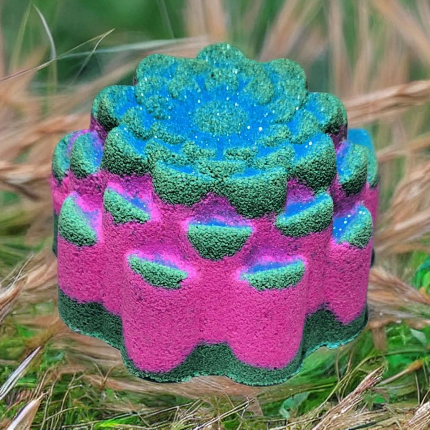 A colorful, flower-shaped Hedgerow Fizzy Bath Bomb from Posh Brats sits on the grass. It features pink, green, and blue layers resembling a succulent with textured petals. The vibrant colors are highlighted by the brown and green grass. This vegan bath bomb enchants with lush essential oils aroma.