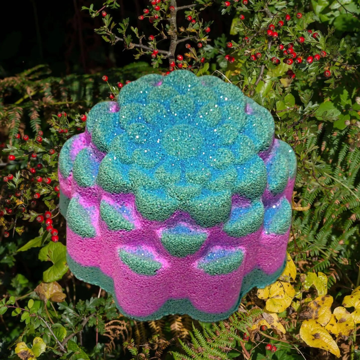 A lively, multicolored sculpture evoking a sea anemone or coral—similar to the Posh Brats Hedgerow Fizzy Bath Bomb with its bright blue and pink colors—rests among green foliage and red berries in a garden.