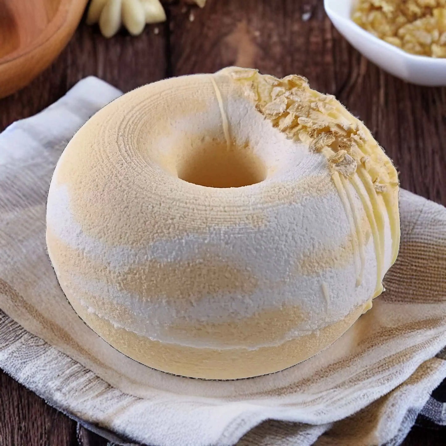A round, creamy Honey Oatmeal Donut Fizzy Bath Bomb by Posh Brats, featuring a light brown and white swirl pattern resembling a doughnut with crushed nuts on top, sits on folded fabric. In the background, theres a bowl of additional crushed nuts.