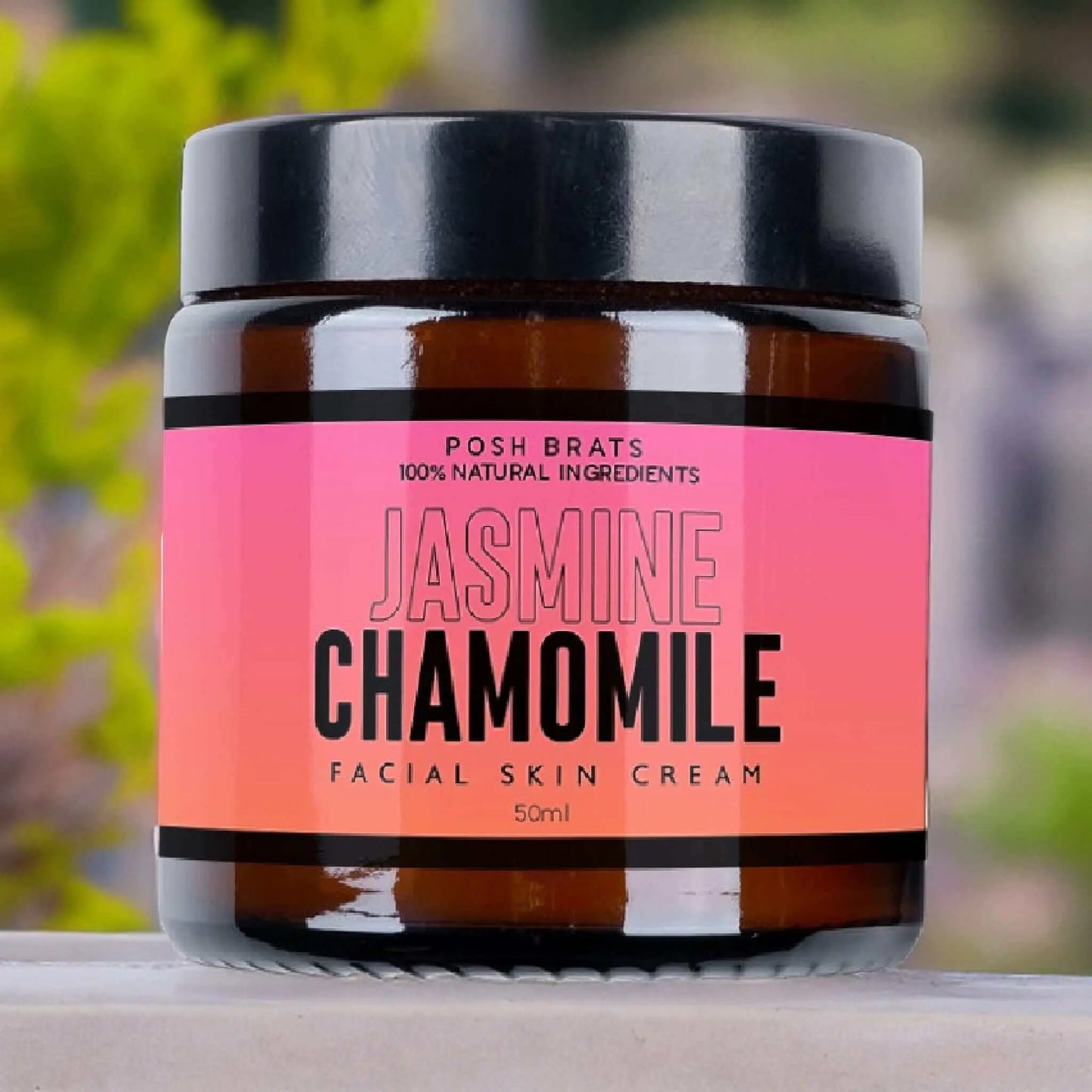 Posh Brats Jasmine Chamomile Skin Cream with a black lid features a pink label reading 100% Natural Ingredients and 50ml. It offers deep hydration and anti-aging benefits, showcased against a softly blurred green and purple backdrop.