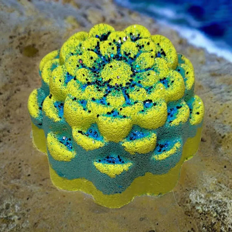 The Aloha Paradise Fizzy Bath Bomb by Posh Brats is a round, textured item resembling a succulent plant. It features yellow and blue hues with black speckles, resting on sandy terrain with a blurred ocean and sky backdrop, capturing the tropical getaway essence.