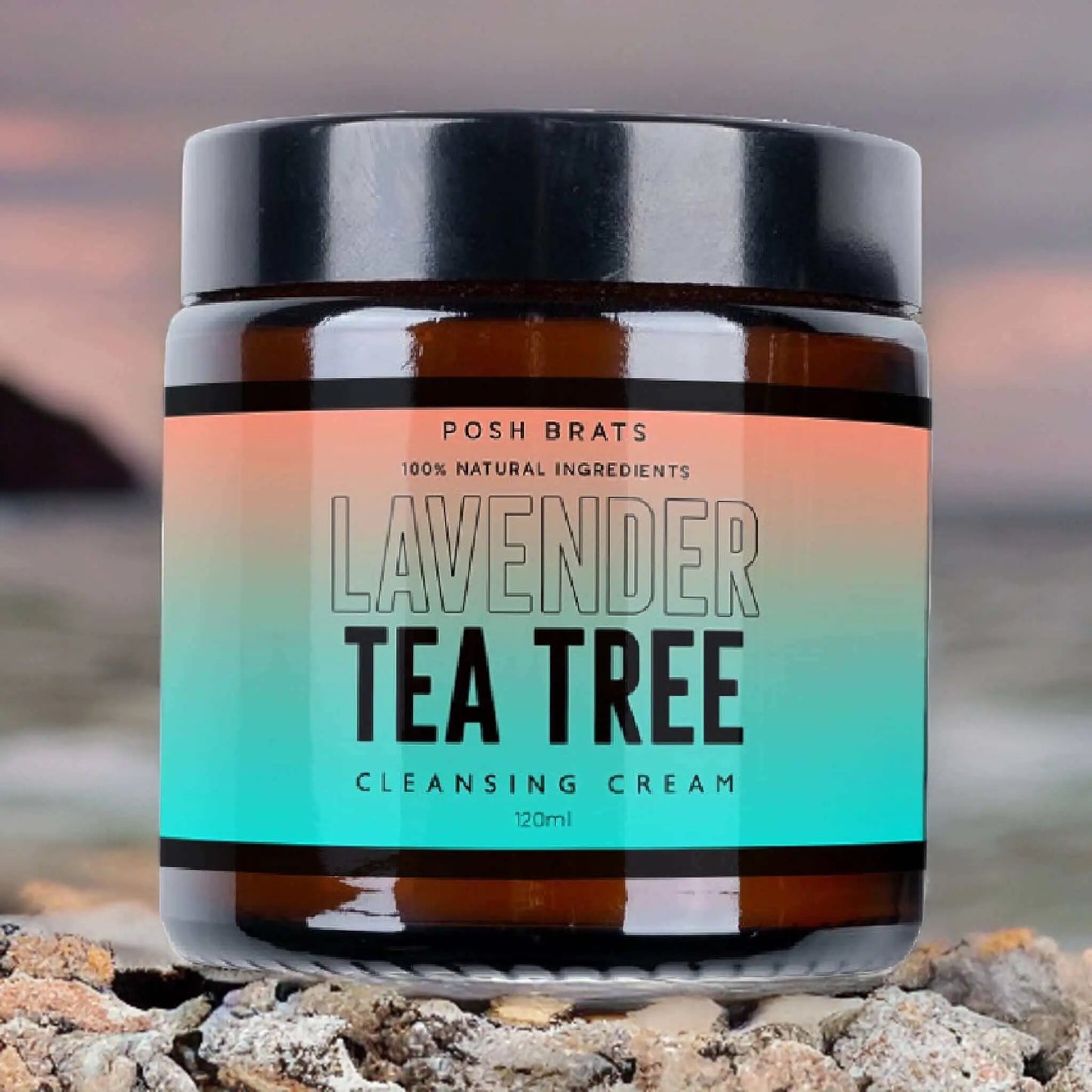 A jar of Posh Brats Lavender Tea Tree Clear Skin Cleansing Cream, with natural essential oils, is placed on rocky terrain before a blurred ocean and cloudy sky. The amber jar has a black lid and colorful gradient label, highlighting its vegan-friendly promise.