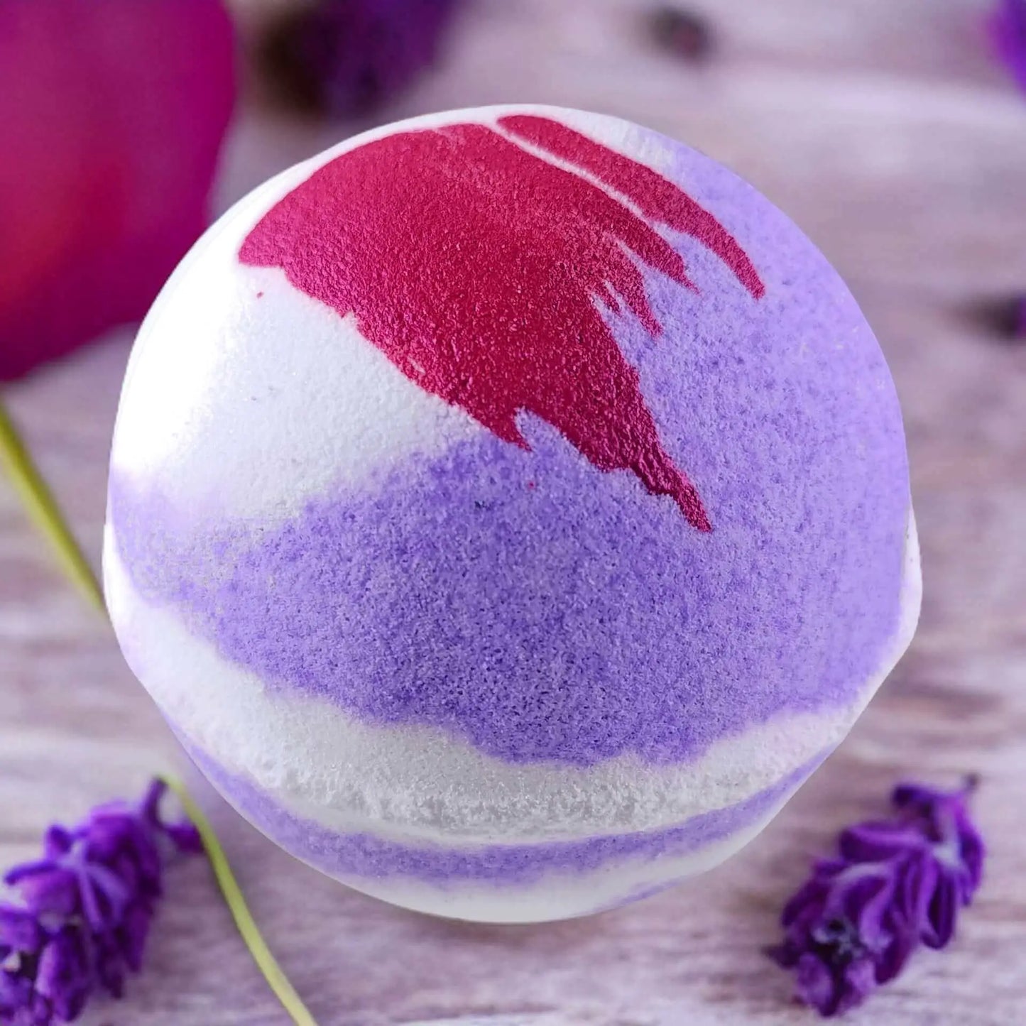 A Posh Brats Luxury Lavender Fizzy Bath Bomb in purple and white with red highlights rests on a wooden surface, surrounded by small purple flowers, delivering a soothing spa-like experience with its calming lavender scent.
