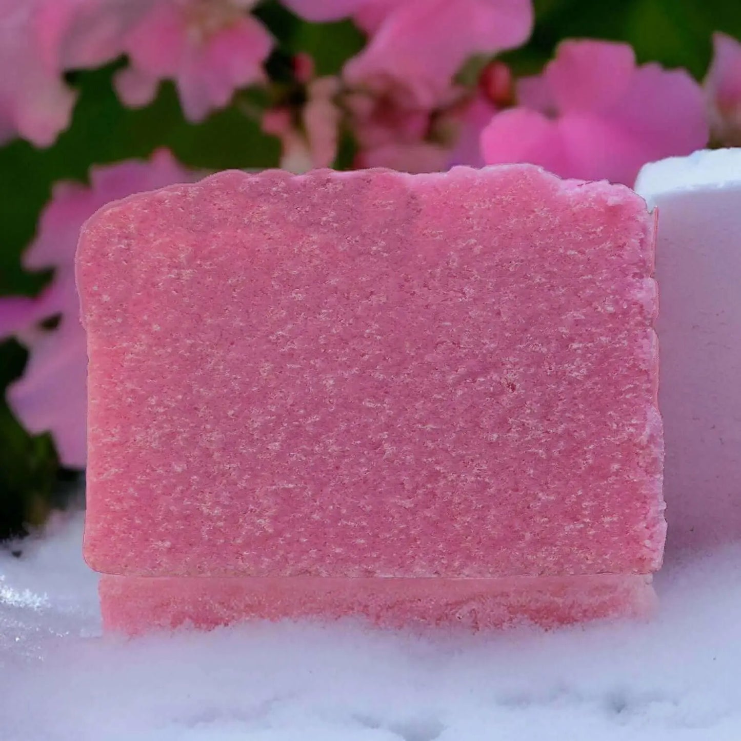 The Marshmallow Magic Sugar Scrub Bar by Posh Brats, featuring a rough texture, is showcased against blurred pink flowers and green leaves, resting on a powder-dusted white surface for an unparalleled exfoliation experience.