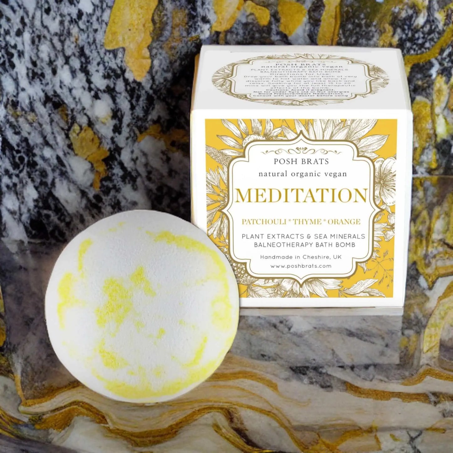 A round bath bomb with yellow accents by Posh Brats rests beside a Meditation Aromatherapy Bath Bomb box. It boasts patchouli, thyme, and orange essential oils with plant extracts and sea minerals for relaxation on the marbled stone surface.