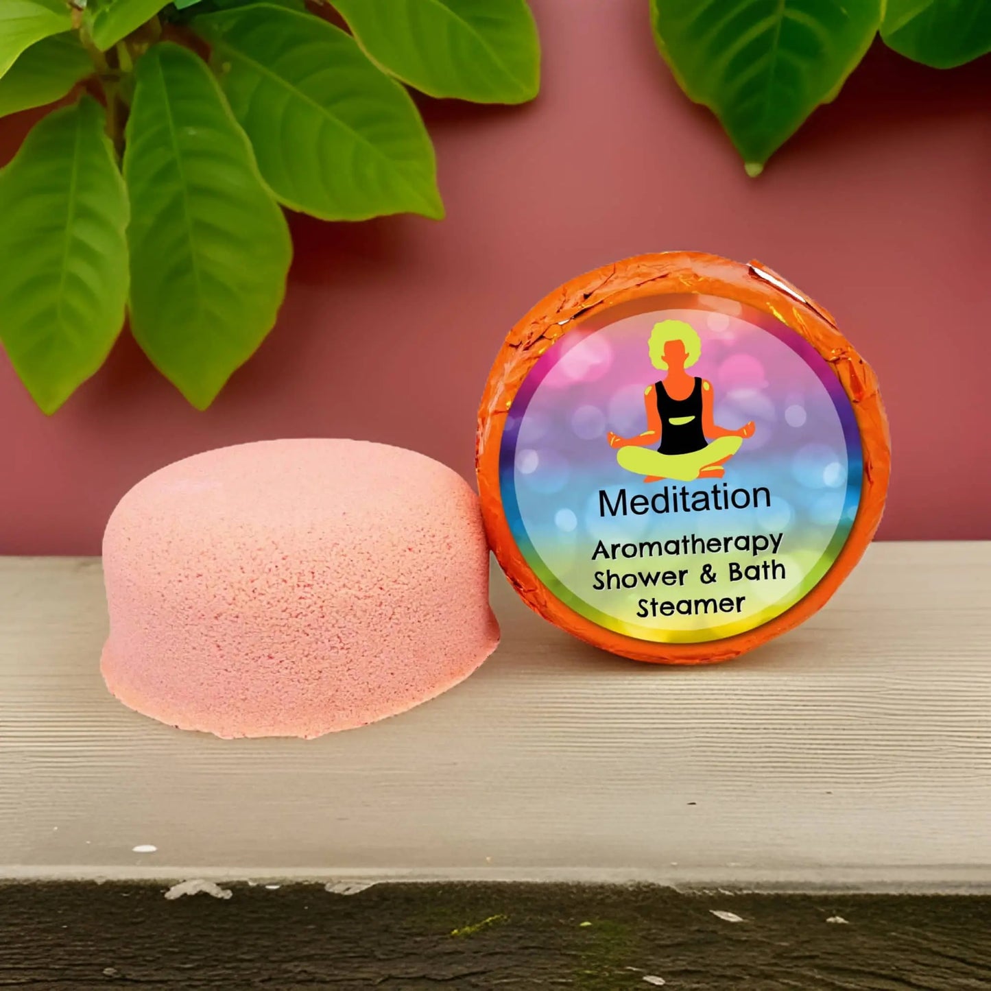 The pink Meditation Aromatherapy Shower Steamer by Posh Brats, infused with essential oils, sits beside its packaging featuring a vibrant meditation label. Green leaves hang against the pink wall, enhancing the tranquil vibe.