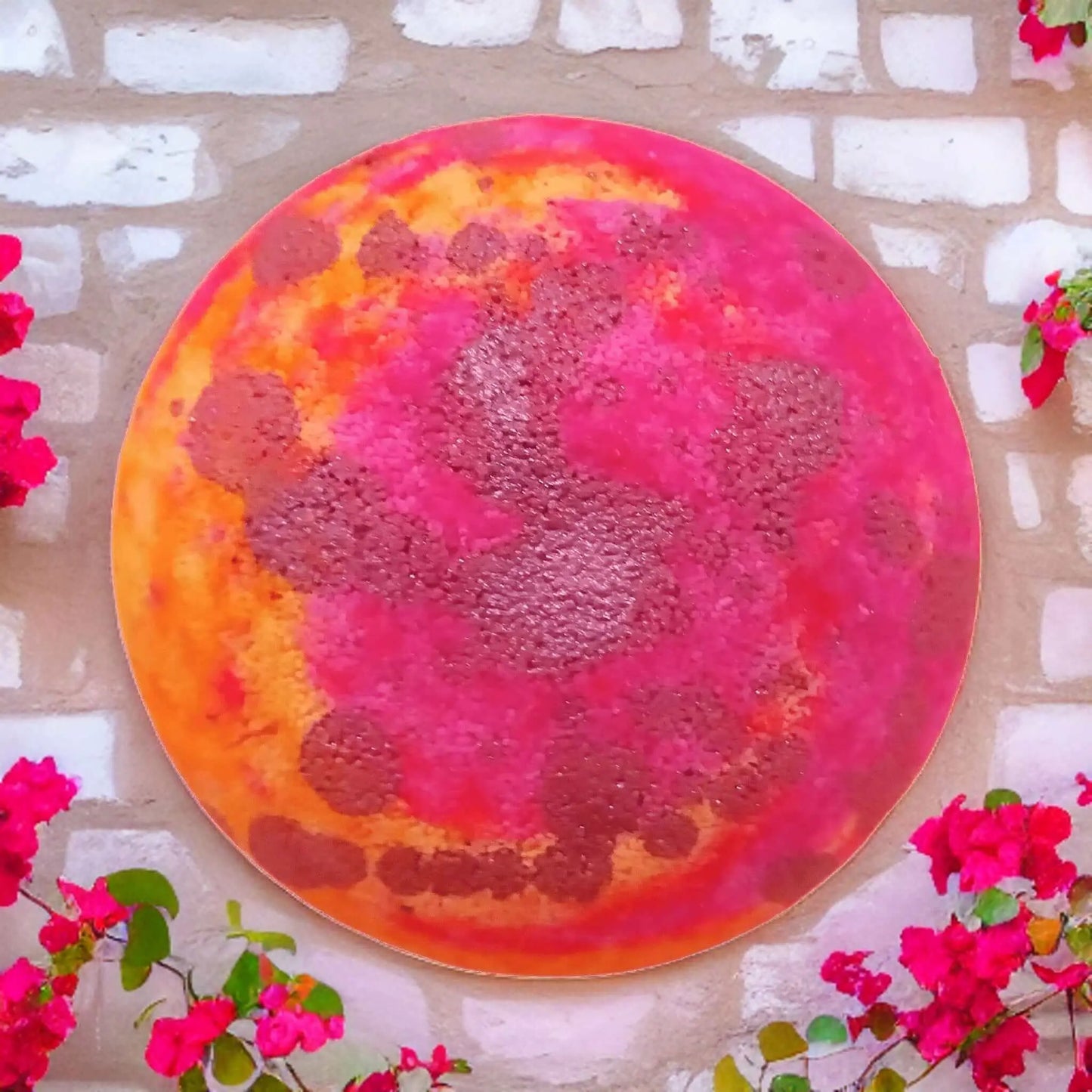 A colorful circular art piece on a brick wall, resembling a planet in pink, orange, and brown hues. It evokes the playful essence of Posh Brats Monkey Farts Fizzy Bath Bomb, framed by vibrant pink flowers on leafy branches contrasting beautifully against the earthy tones of the wall.