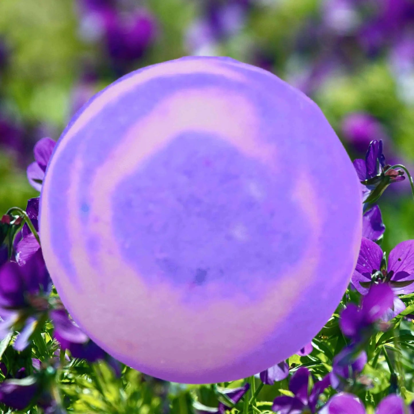 A Victorian Parma Violet Fizzy Bath Bomb by Posh Brats, with its lavender and pink hues, rests among vibrant purple flowers and lush green leaves, its soft effervescence echoing the delicacy of a soap bubble against the rich colors of the surrounding flora.