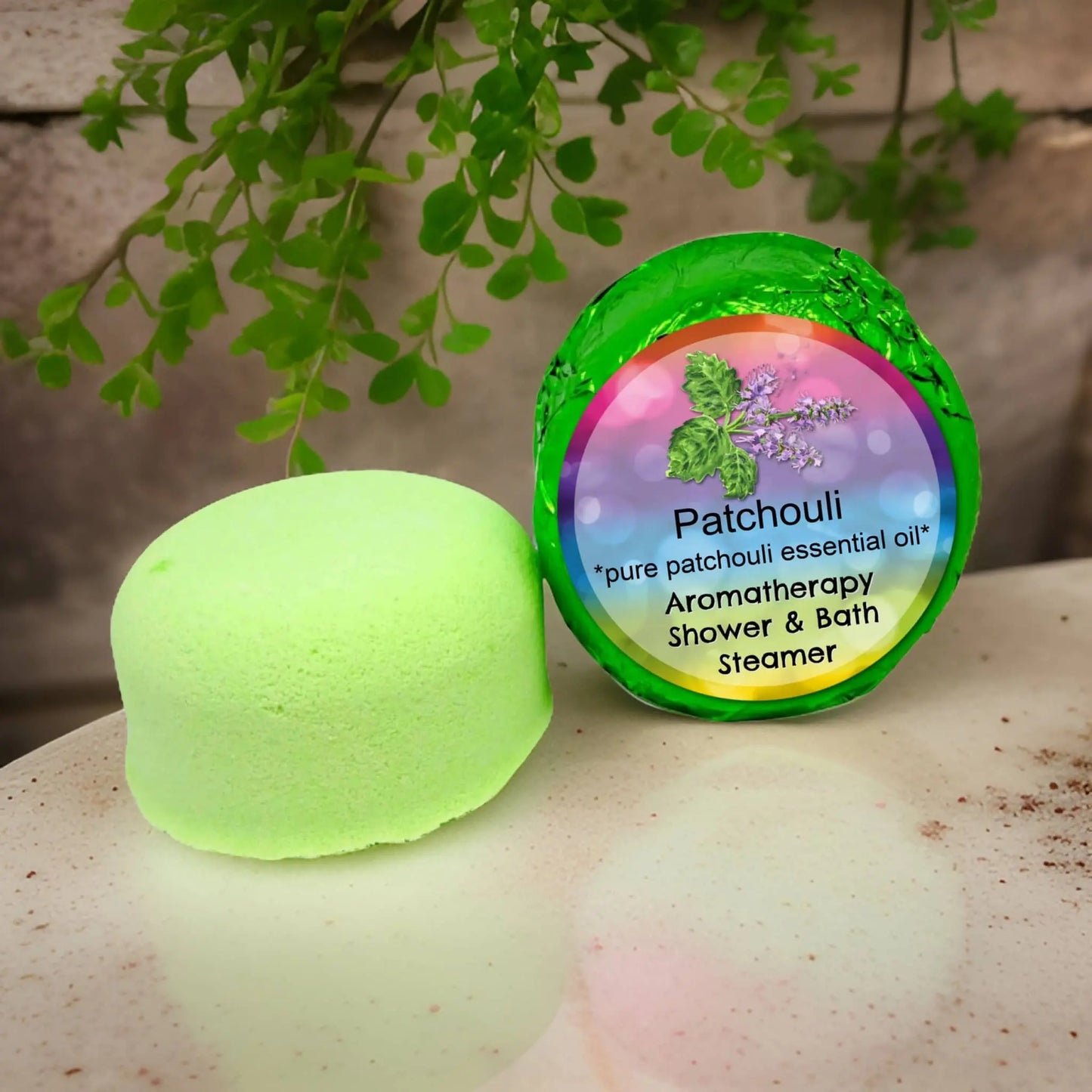 A Posh Brats Patchouli Aromatherapy Shower Steamer with bright green packaging, featuring patchouli leaves and essential oils, rests on a countertop beside an unfolded piece amid lush green foliage.