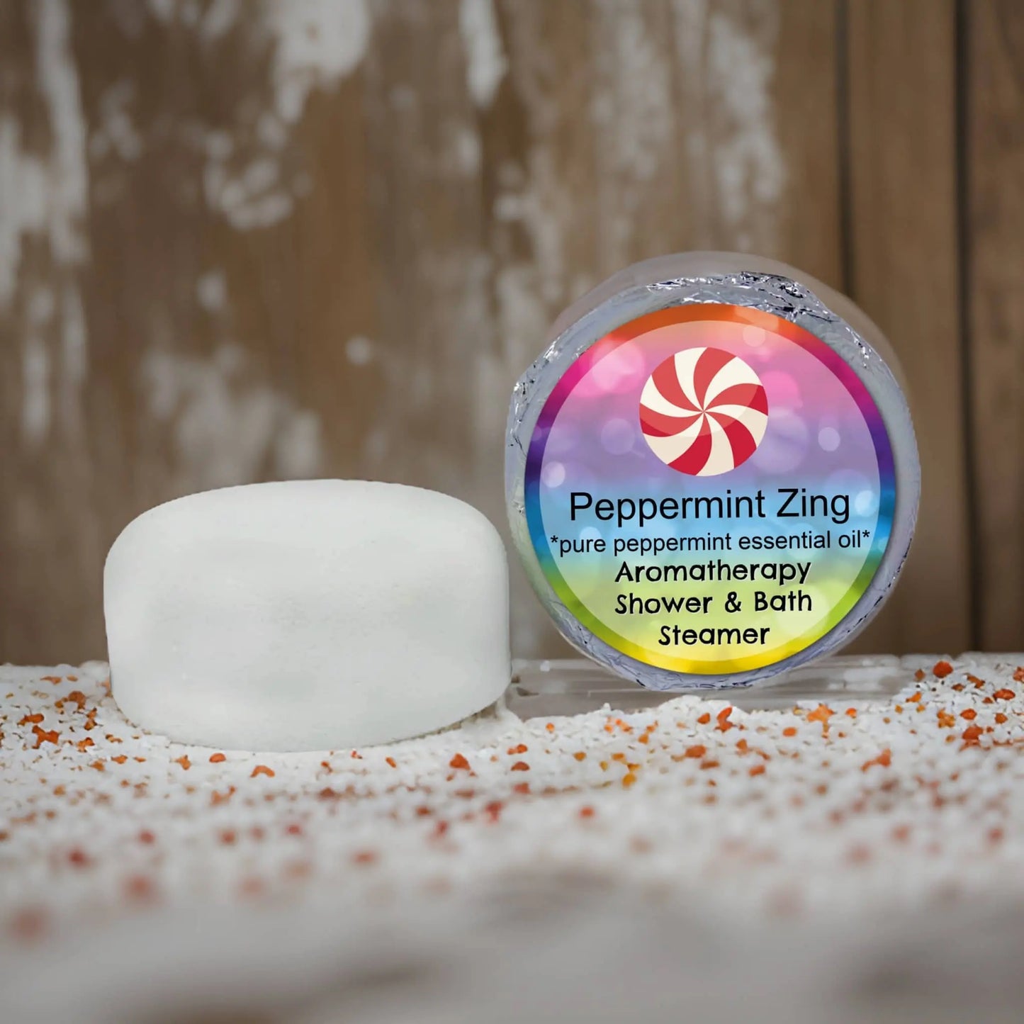 A round, white bath steamer sits next to its colorful packaging featuring a red and white swirl. The label reads Peppermint Zing by Posh Brats, packed with natural essential oils for an invigorating aromatherapy experience in your shower.