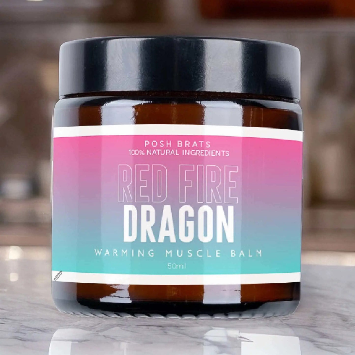 A jar of Posh Brats Red Dragon Warming Balm Aromatherapy features a gradient pink and blue label with bold white text, emphasizing its natural ingredients. It rests on a reflective surface, offering effective heat therapy with elegance against a blurred background.