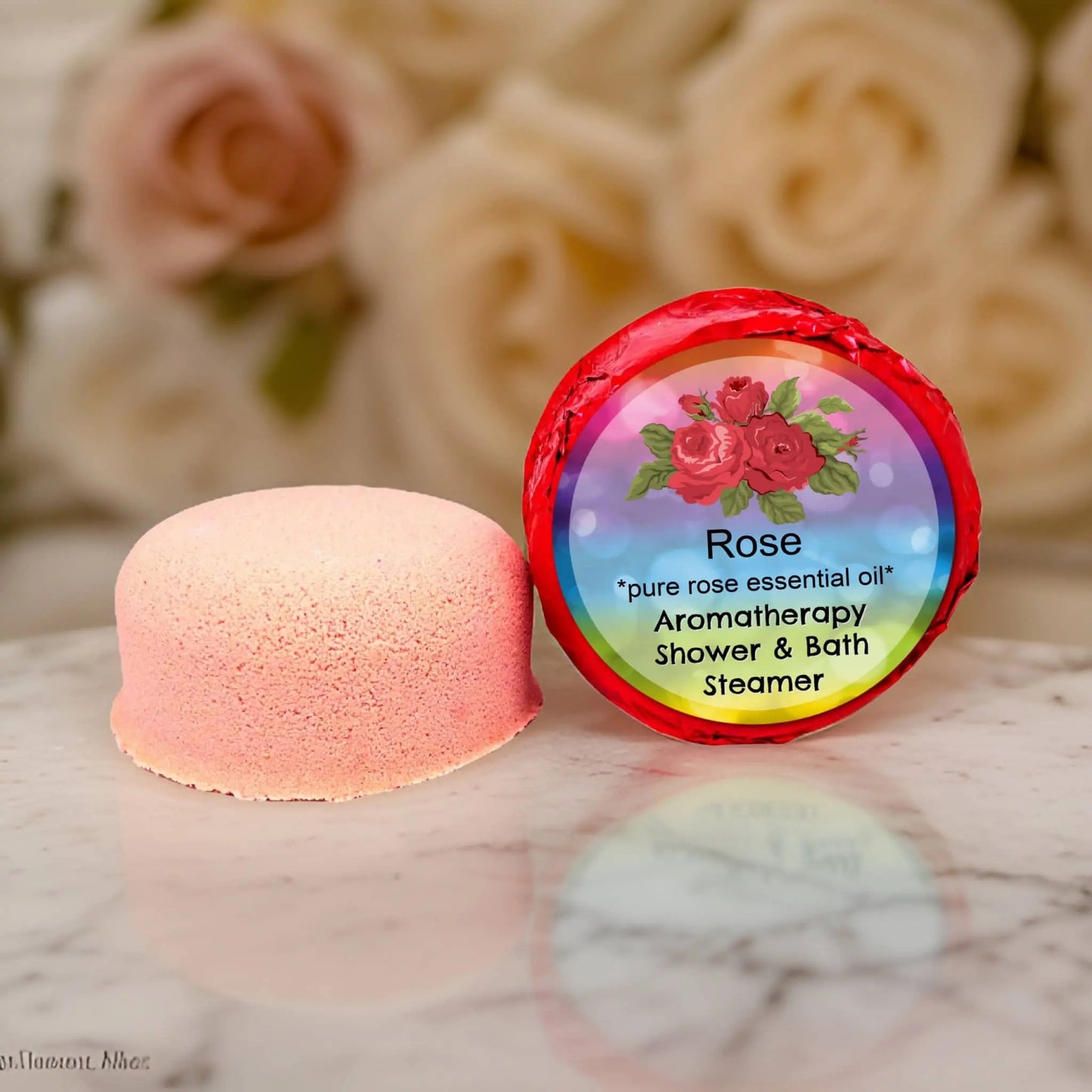 A pink Rose Aromatherapy Shower Steamer by Posh Brats is displayed on a marble surface near its vibrant, rose-themed packaging. Cream and pink roses blur in the background, as the package highlights a spa-like experience using pure rose essential oils.