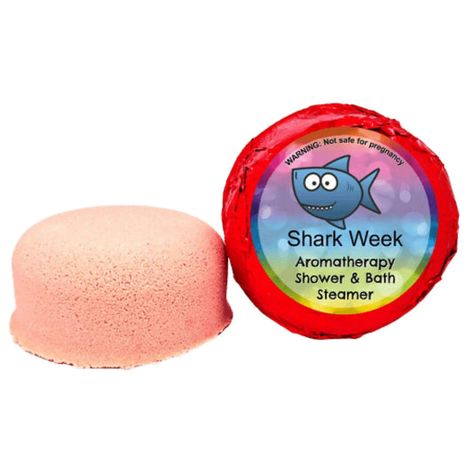 A pink shower steamer infused with essential oils sits next to its red wrapper labeled Shark Week PMS Aromatherapy Shower Steamer by Posh Brats, featuring a cartoon shark and a warning that its not safe for pregnancy.