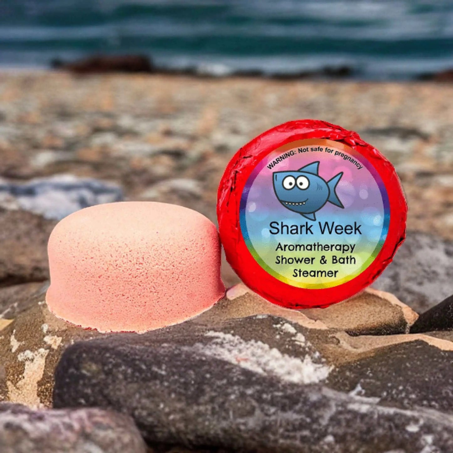A round, peach bath steamer enriched with essential oils rests on a rocky beach beside its packaging, which displays a smiling shark and reads Shark Week PMS Aromatherapy Shower Steamer by Posh Brats. The soothing ocean forms the distant backdrop.