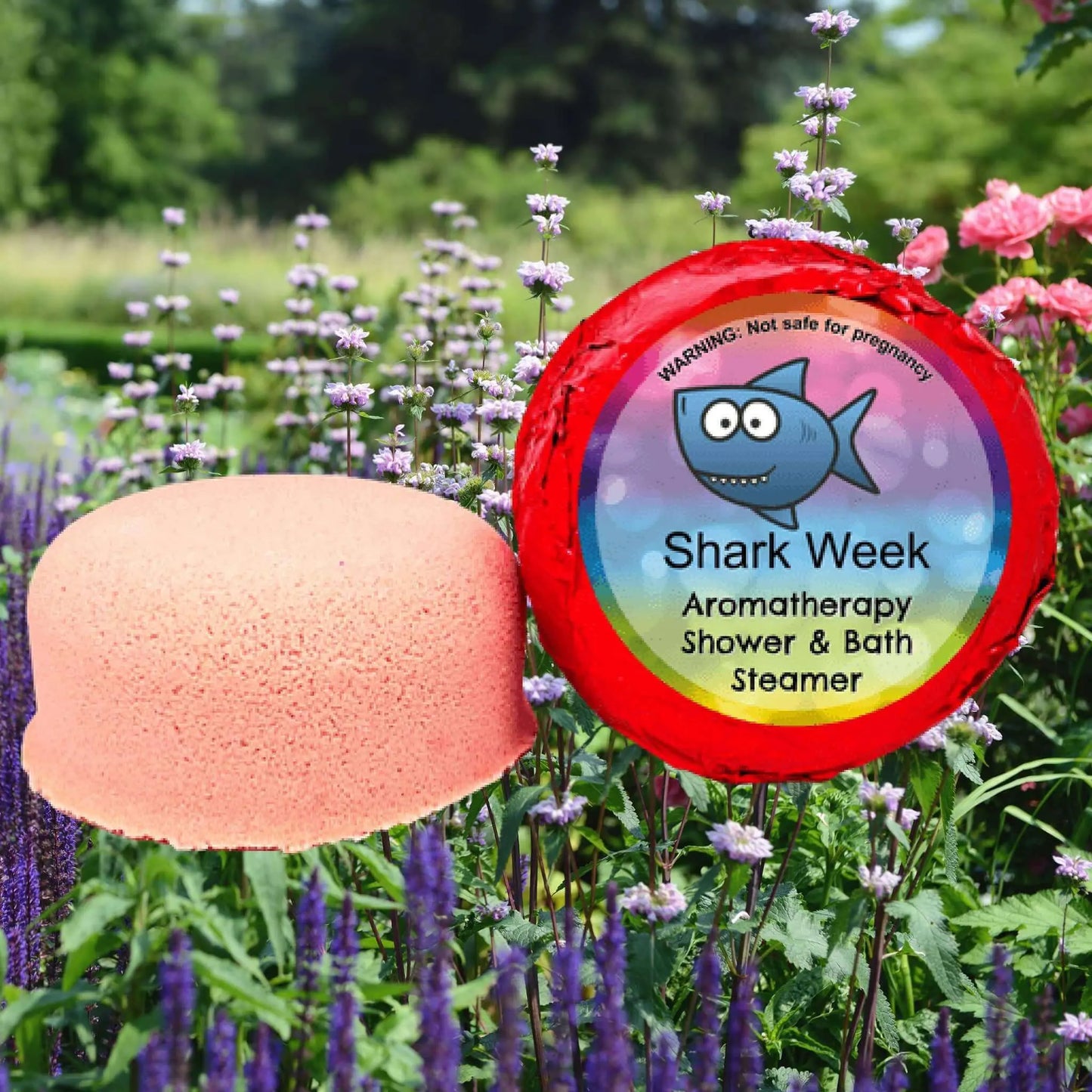 The Posh Brats Shark Week PMS Aromatherapy Shower Steamer, round and pink with a colorful label featuring a cartoon shark, sits against a vibrant garden backdrop of purple and pink flowers. The label emphasizes its essential oil blend for an ideal aromatherapy session.