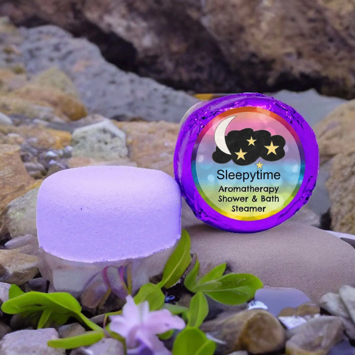 A Posh Brats purple Sleepytime Aromatherapy Shower Steamer, infused with lavender and chamomile, rests among rocks, small green plants, and a purple flower. The moon and stars packaging enhances its nighttime theme with natural essential oils.
