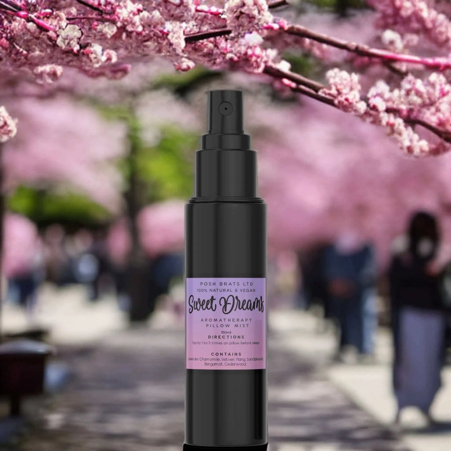 A focused black bottle labeled Sweet Dreams Magnesium Aromatherapy Pillow Mist by Posh Brats is nestled against a dreamy blur of blooming cherry blossoms and a pathway with people strolling, inviting relaxation and restful nights.