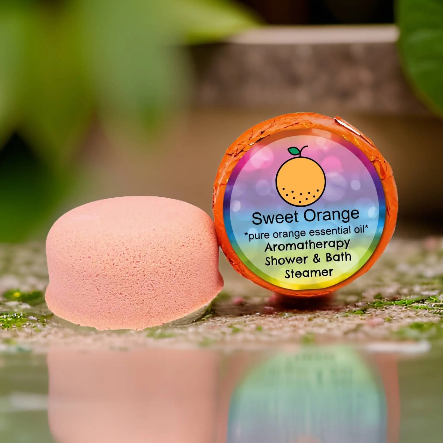 A pink shower and bath steamer labeled Sweet Orange Aromatherapy Shower Steamer by Posh Brats boasts a vibrant wrapper. It contains pure essential oil, resting on a reflective surface and surrounded by lush green foliage.