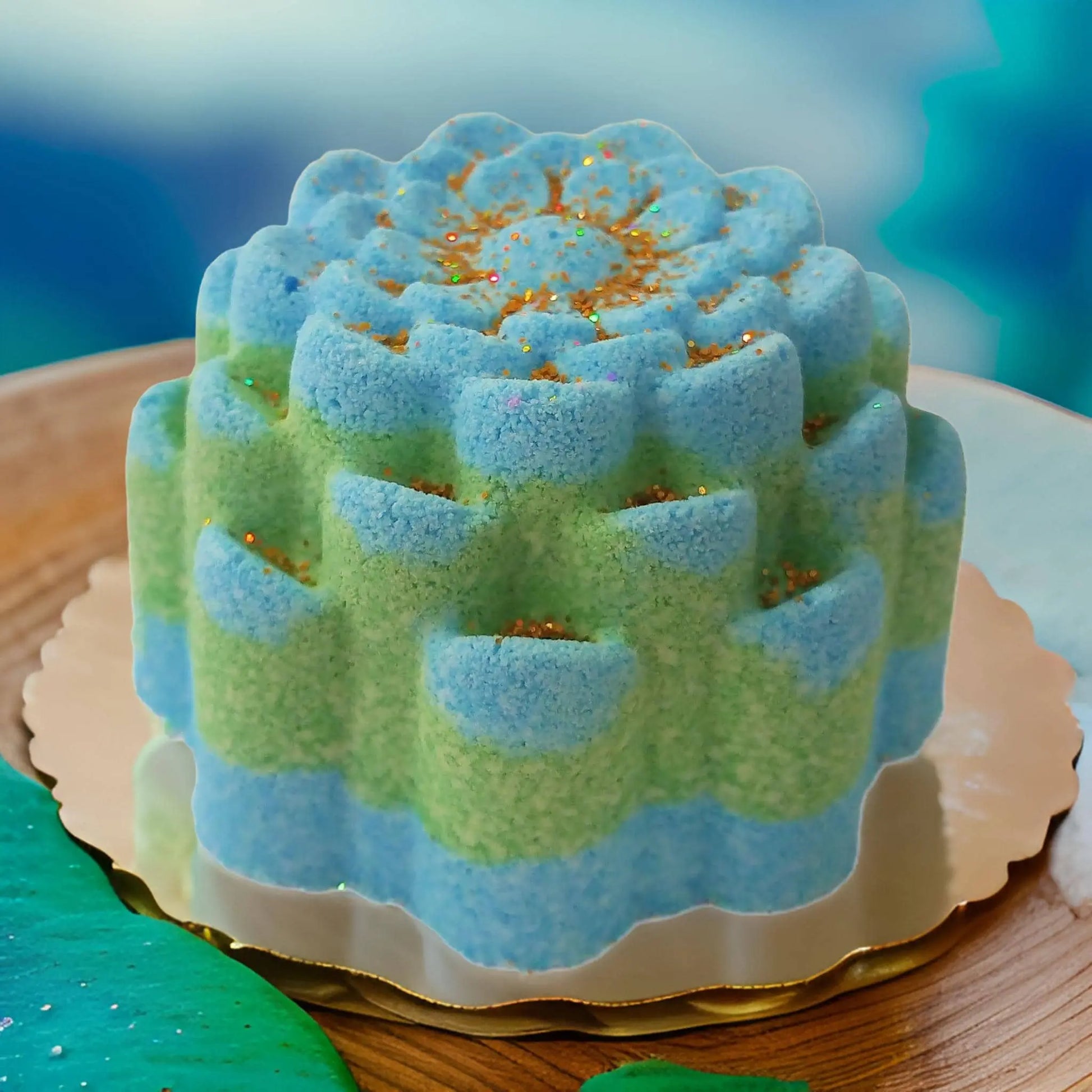 A blue and green mousse cake with a floral pattern on a gold base resembles Posh Brats Ocean Breeze Fizzy Bath Bomb. Decorated with gold sprinkles, it features a smooth texture and intricate blooming flower design. The softly blurred background highlights its vibrant allure.