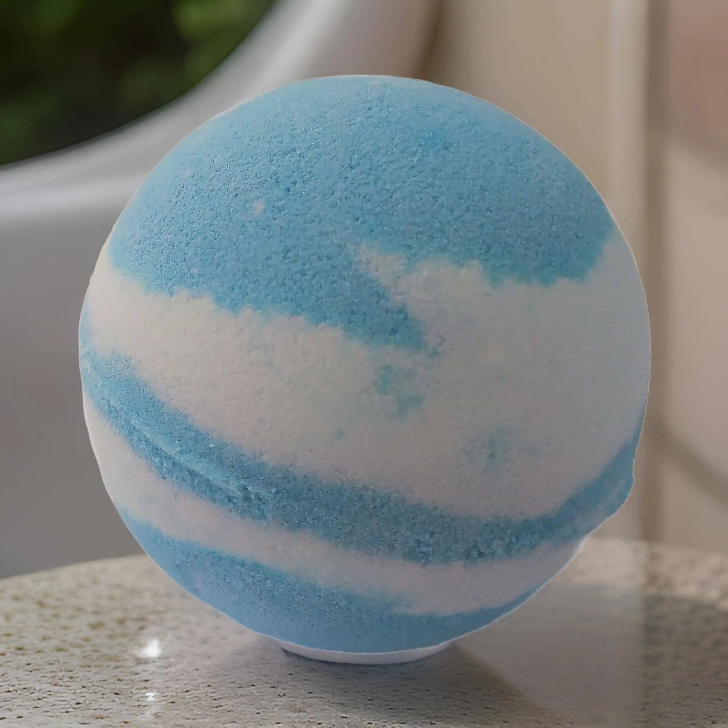 The Peony & Cotton Blossom Fizzy Bath Bomb by Posh Brats, with nourishing ingredients, features swirling blue and white hues on a textured surface. Its blurred backdrop with greenery evokes a luxurious bathroom retreat.