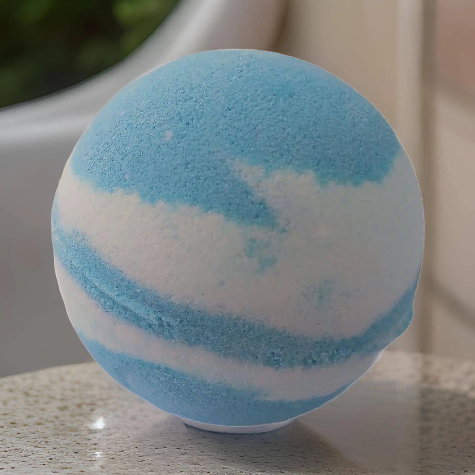 The Peony & Cotton Blossom Fizzy Bath Bomb by Posh Brats, with nourishing ingredients, features swirling blue and white hues on a textured surface. Its blurred backdrop with greenery evokes a luxurious bathroom retreat.