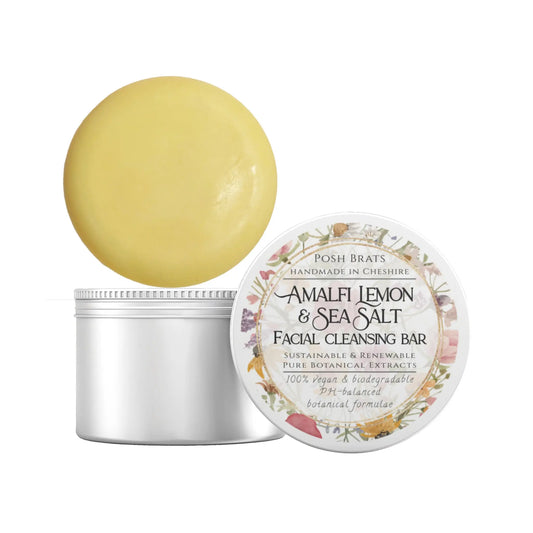 A round, yellow facial cleansing bar lies next to its open container. The lid features floral designs and reads Amalfi Lemon + Sea Salt Cleansing Bar by Posh Brats, highlighting its natural and vegan ingredients.