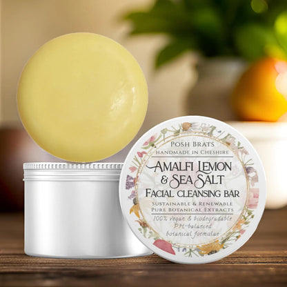 The image showcases a Posh Brats Amalfi Lemon + Sea Salt Cleansing Bar on an open container, its label highlighting sustainable, vegan ingredients. A softly blurred background with warm tones and greenery enhances the products natural appeal.