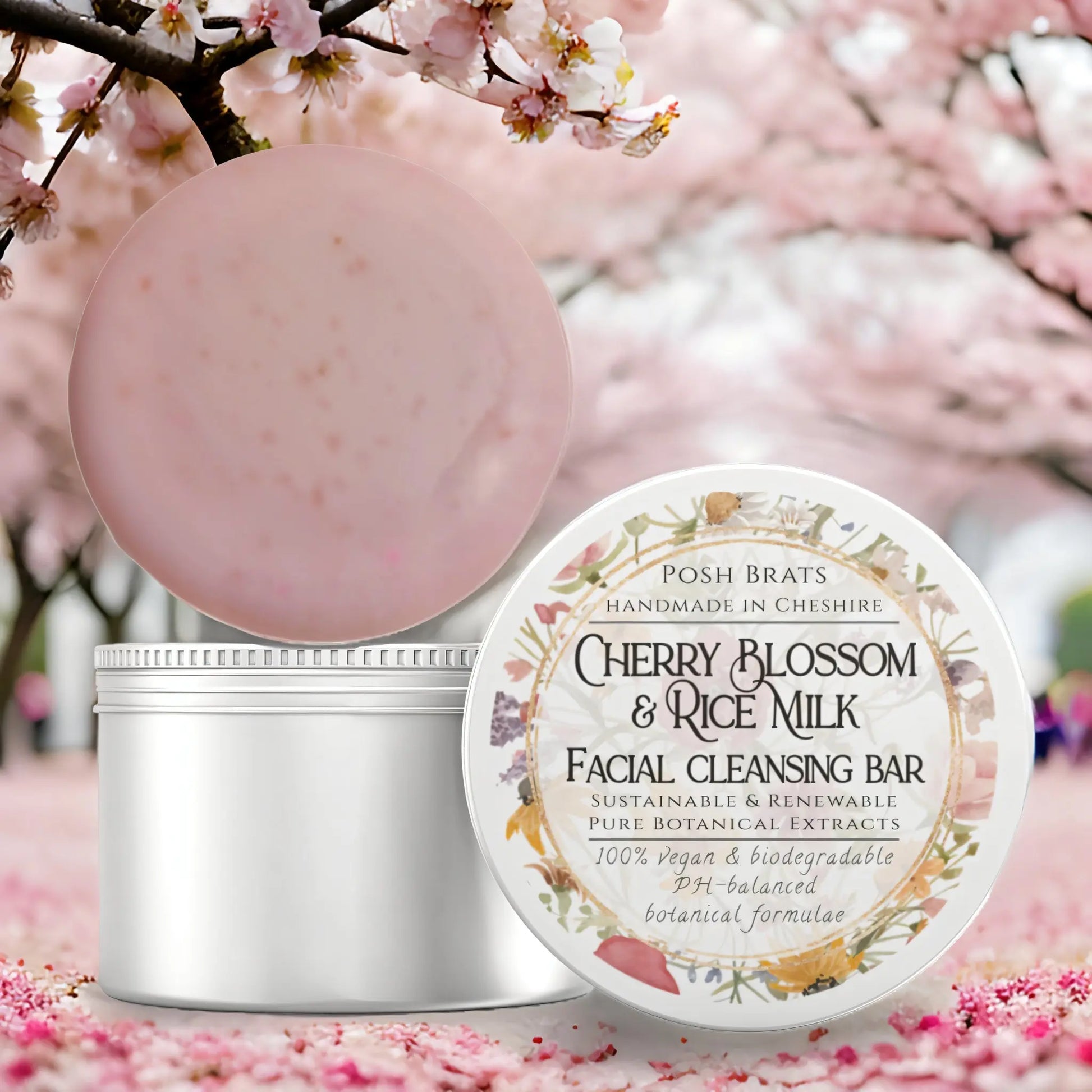 An open circular tin labeled Cherry Blossom + Rice Milk Cleansing Bar by Posh Brats shows a pink bar beside cherry blossoms. This vegan and biodegradable product ensures a gentle and refreshing cleanse.