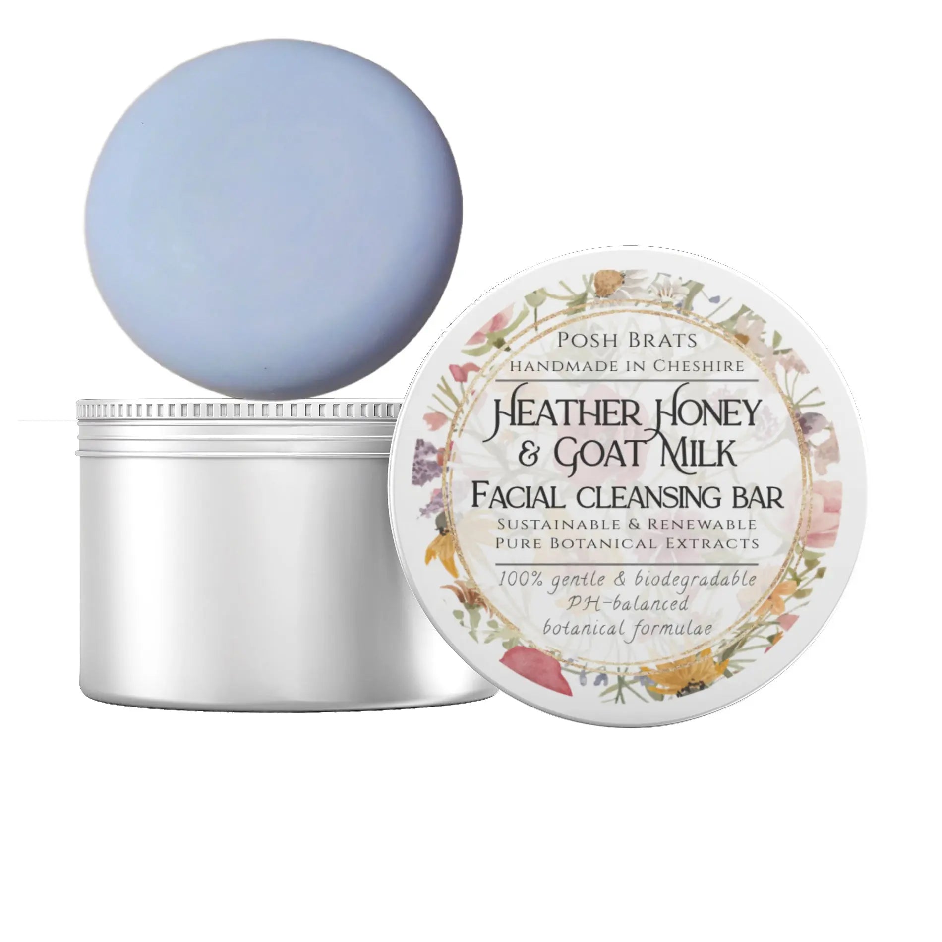 A round, light blue Posh Brats facial cleansing bar rests on a partially opened white jar. The lid features a floral label stating Heather Honey + Goat Milk Facial Cleansing Bar, emphasizing its gentle, biodegradable, plant-based ingredients.