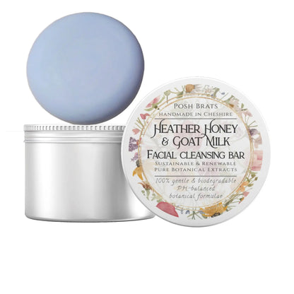 A round, light blue Posh Brats facial cleansing bar rests on a partially opened white jar. The lid features a floral label stating Heather Honey + Goat Milk Facial Cleansing Bar, emphasizing its gentle, biodegradable, plant-based ingredients.