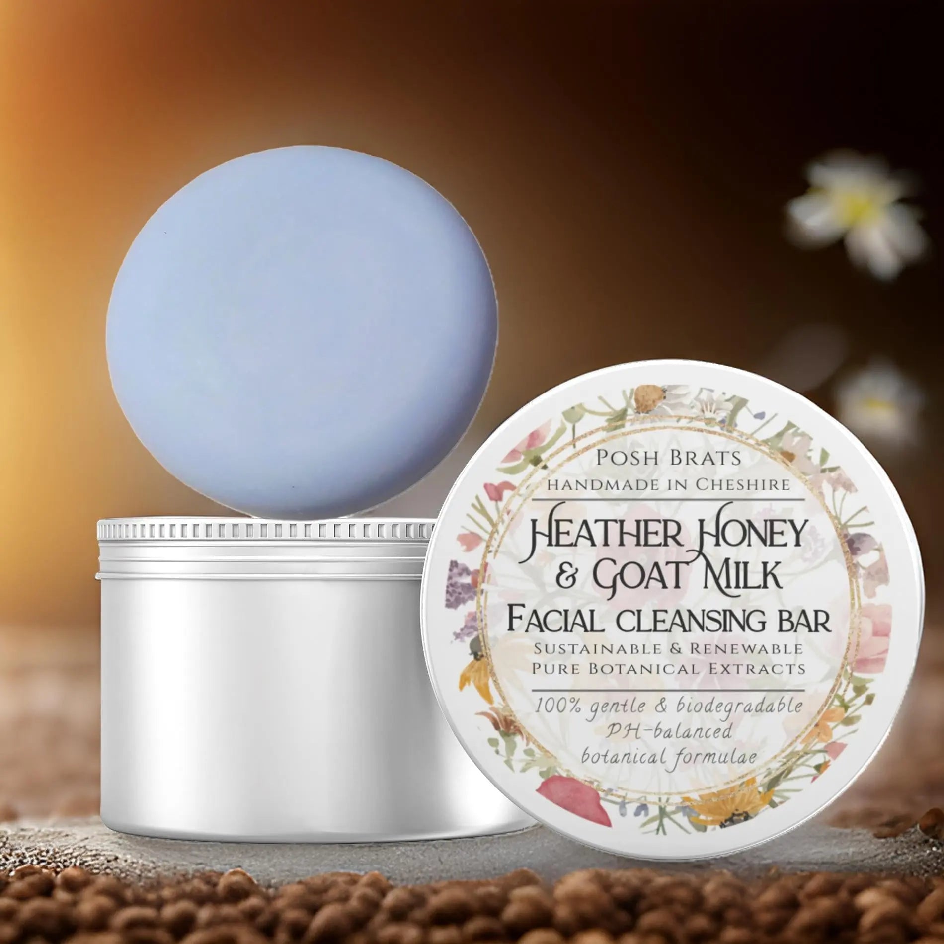 A Posh Brats Heather Honey + Goat Milk Facial Cleansing Bar sits on an open white container, with soft-focus flowers gently in the background.