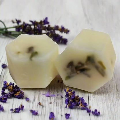 Two hexagonal English Lavender Botanical Butter Bath Melts by Posh Brats, enriched with shea butter and lavender buds, rest on a wooden surface. Fresh English lavender sprigs and scattered petals complete the natural, calming setting.