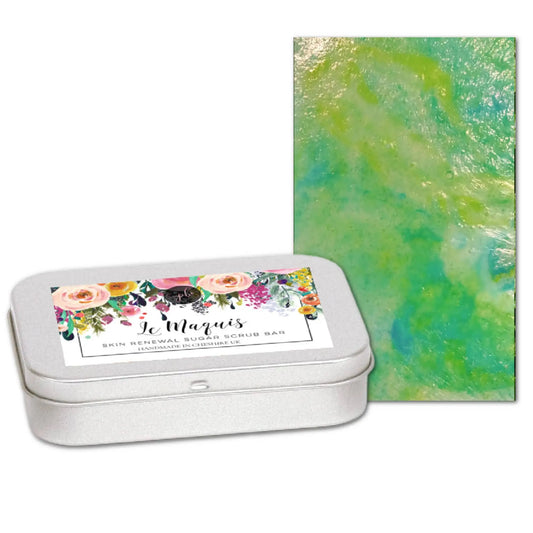 A silver tin labeled Le Macaron Skin Renewal Sugar Scrub Bar features a vibrant floral design with a Mediterranean scent. Next to it is the luxurious Le Maquis Corsican Maquis Sugar Scrub Bar by Posh Brats, boasting green and yellow swirls inspired by Corsican plant extracts.