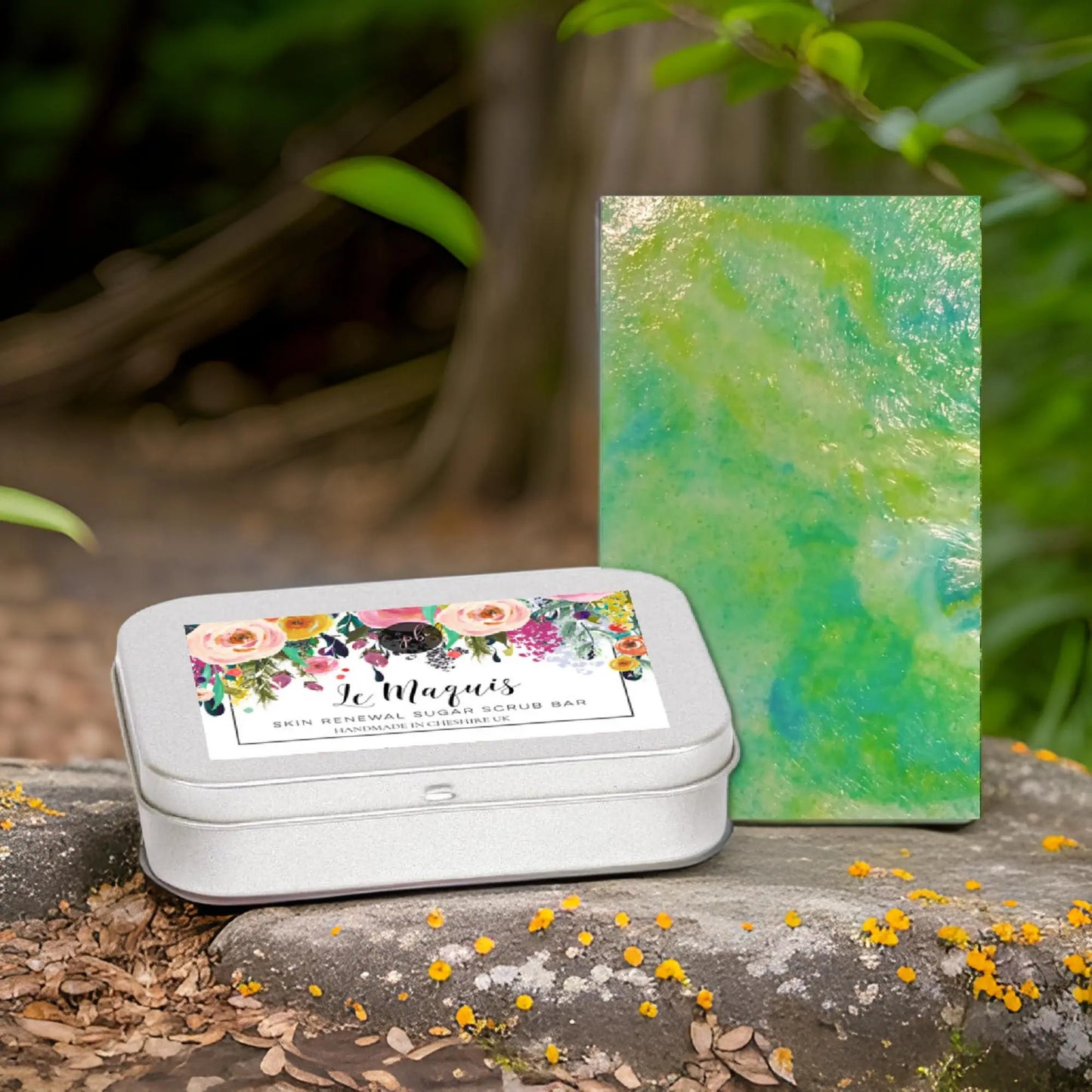 A silver tin labeled Le Maquis Corsican Maquis Sugar Scrub Bar from Posh Brats, with floral designs, rests on stone, exuding a Mediterranean scent. Next to it is a green and blue marbled bar. The backdrop shows lush greenery, scattered yellow flowers, and hints of Corsican plant extracts.