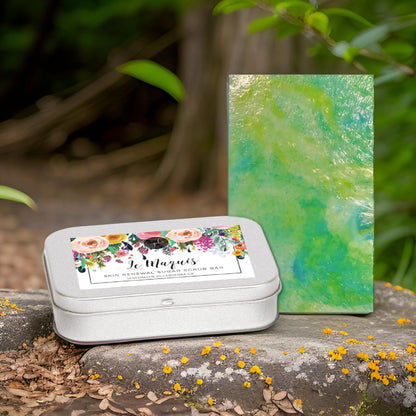 A silver tin labeled Le Maquis Corsican Maquis Sugar Scrub Bar from Posh Brats, with floral designs, rests on stone, exuding a Mediterranean scent. Next to it is a green and blue marbled bar. The backdrop shows lush greenery, scattered yellow flowers, and hints of Corsican plant extracts.