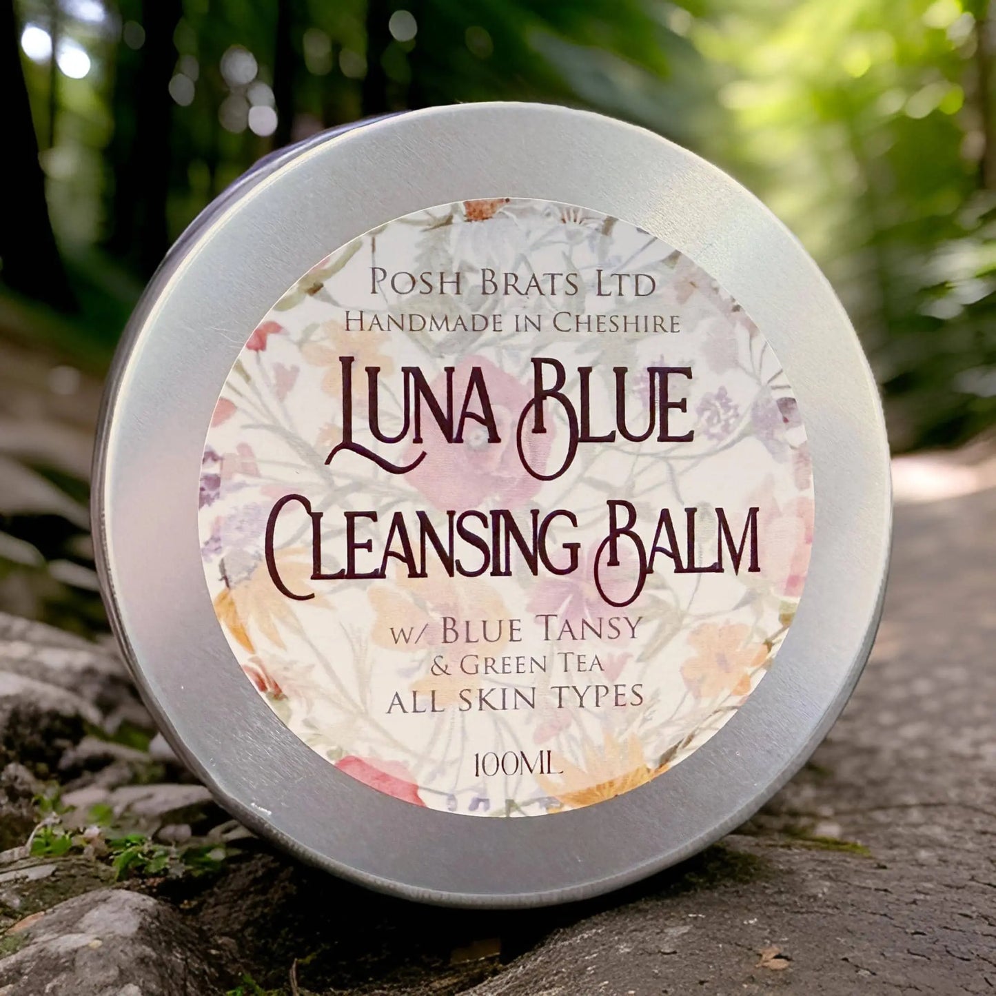 A round silver tin of Posh Brats Luna Blue Tansy Cleansing Balm with a floral design sits outdoors on stone, promoting blue tansy oil and green tea for radiant skin, suitable for all types.