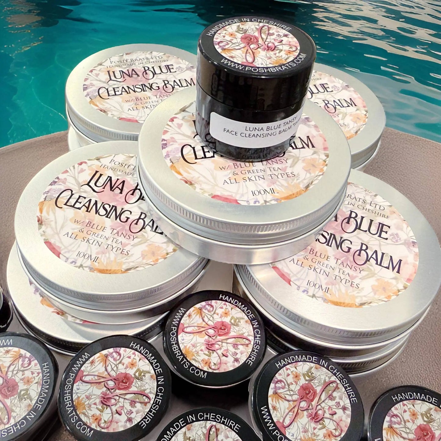 Posh Brats Luna Blue Tansy Cleansing Balm tins and jars, adorned with floral lids, are elegantly displayed by a pool. The balm is infused with blue tansy oil, suitable for all skin types, and promises radiant skin. Handmade in Cheshire.