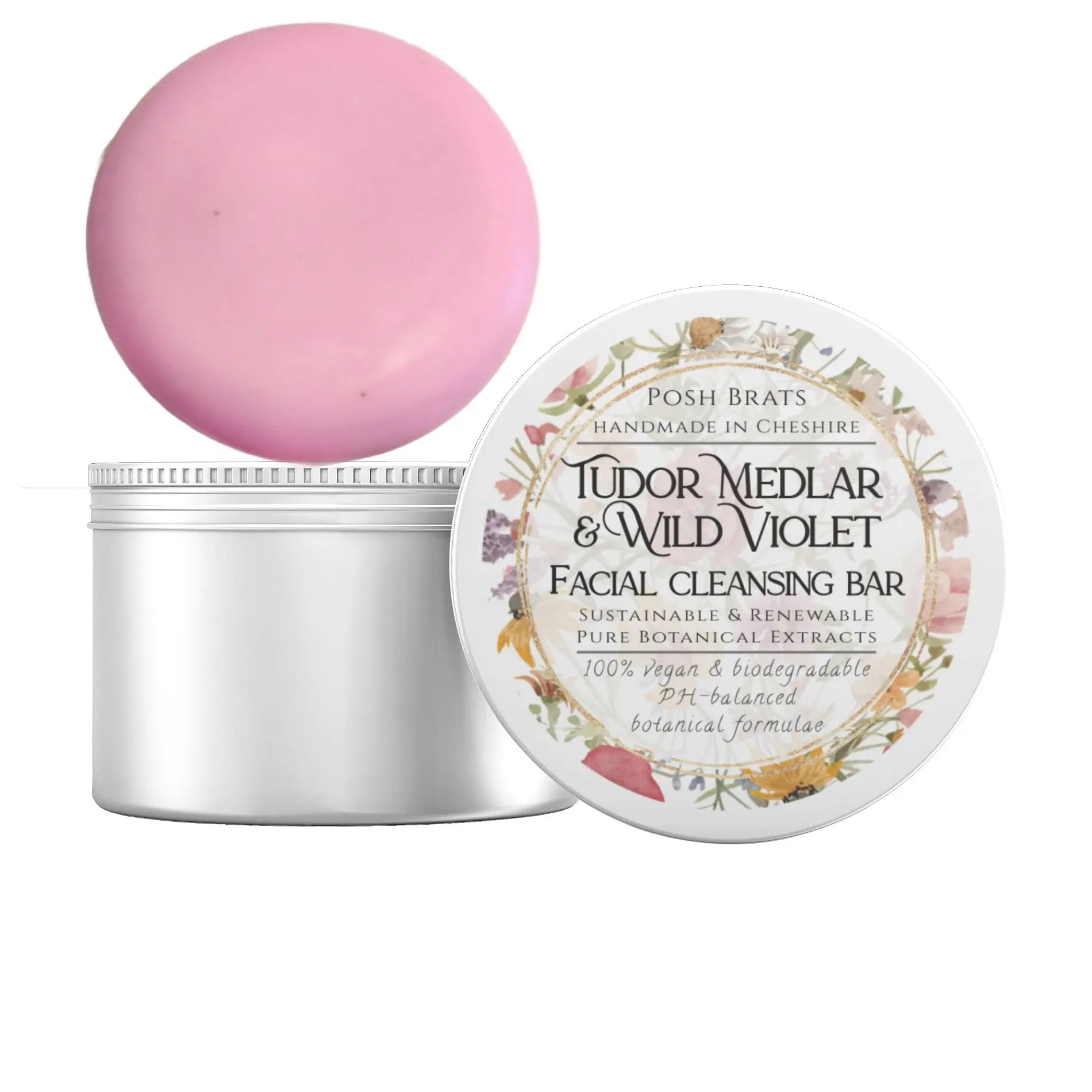 The round, pink Tudor Medlar + Wild Violet Cleansing Bar by Posh Brats sits in a white tin adorned with botanical designs, featuring wild rose notes. Its 100% vegan, biodegradable, and pH-balanced.