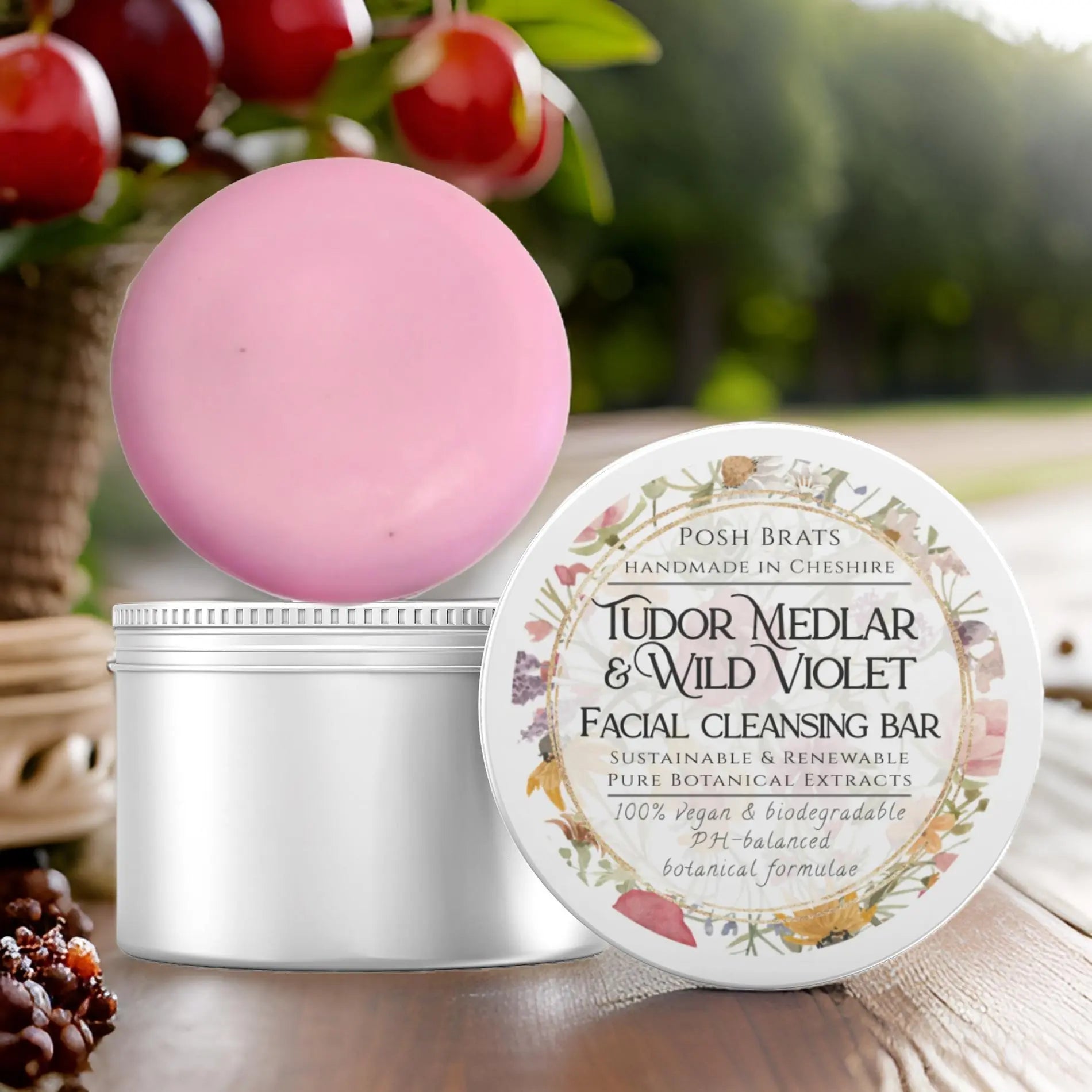 A pink Tudor Medlar + Wild Violet Cleansing Bar by Posh Brats, with an open lid showcasing its floral label, rests among cherries and lush greenery, hinting at wild rose elegance.