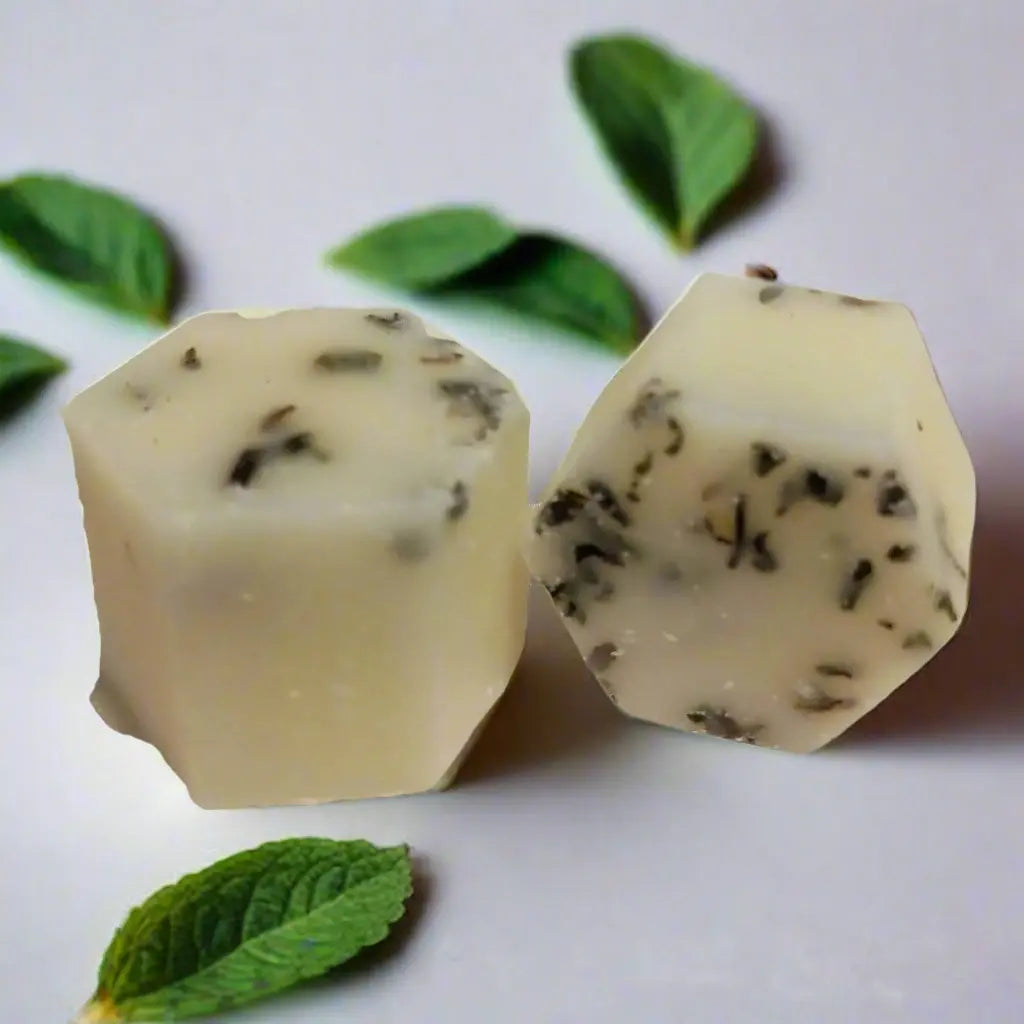 Two hexagonal pieces of white fudge with herb flecks rest on a white surface, surrounded by fresh mint leaves, evocative of the soothing aroma and creamy texture of Posh Brats Peppermint Botanical Butter Bath Melt.