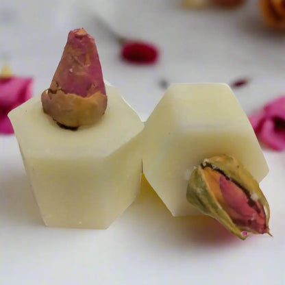 Two hexagonal Damask Rose Botanical Butter Bath Melts from Posh Brats, featuring dried rosebuds on top, rest on a white surface. Blurred petals in the background accentuate the botanical butter look.