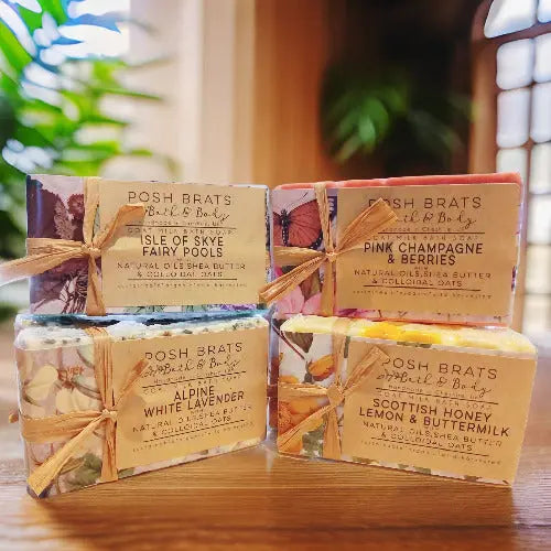 Four Cold Process Castille Soap Bars from Posh Brats, boasting scents like Isle of Skye Fairy Pools, Alpine White Lavender, Pink Champagne & Berries, and Scottish Honey Lemon & Buttermilk, are stacked against a blurred background of green plants and a window.