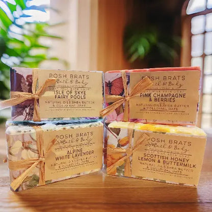 Four Cold Process Castille Soap Bars from Posh Brats, boasting scents like Isle of Skye Fairy Pools, Alpine White Lavender, Pink Champagne & Berries, and Scottish Honey Lemon & Buttermilk, are stacked against a blurred background of green plants and a window.