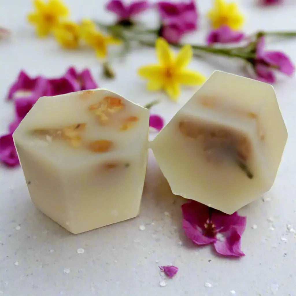 Two hexagonal Woodbine Jessamy Botanical Butter Bath Melts by Posh Brats, white and infused with cocoa butter and floral pieces, rest gracefully amid scattered yellow and pink flower petals.