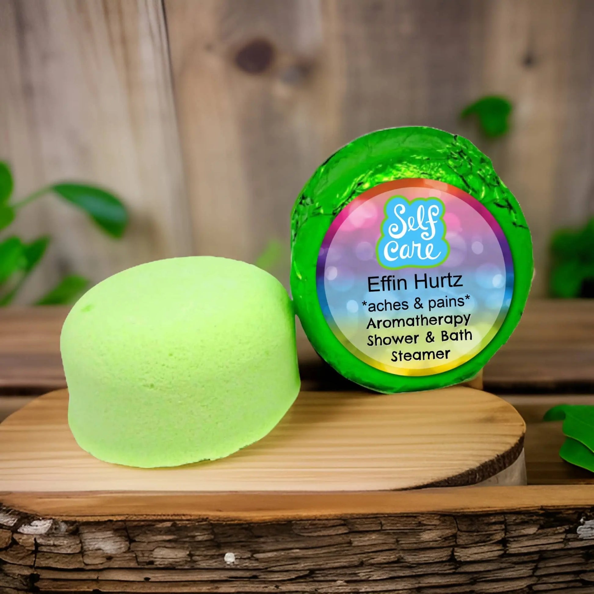 On a wooden surface lies a round, green Posh Brats Effin Hurtz Aromatherapy Shower Steamer with packaging labeled Self Care, aches & pains, set against green leaves with hints of Lavender Marjoram essential oils for a soothing experience.