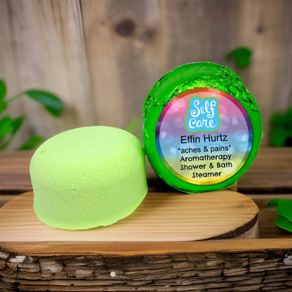 On a wooden surface lies a round, green Posh Brats Effin Hurtz Aromatherapy Shower Steamer with packaging labeled Self Care, aches & pains, set against green leaves with hints of Lavender Marjoram essential oils for a soothing experience.