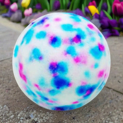 A ball with a white base, speckled in blue, pink, and purple like the Baby Powder Fizzy Bath Bomb by Posh Brats, rests on a paved path. In the background, vibrant tulips and lush greenery form a soothing foamy paradise.